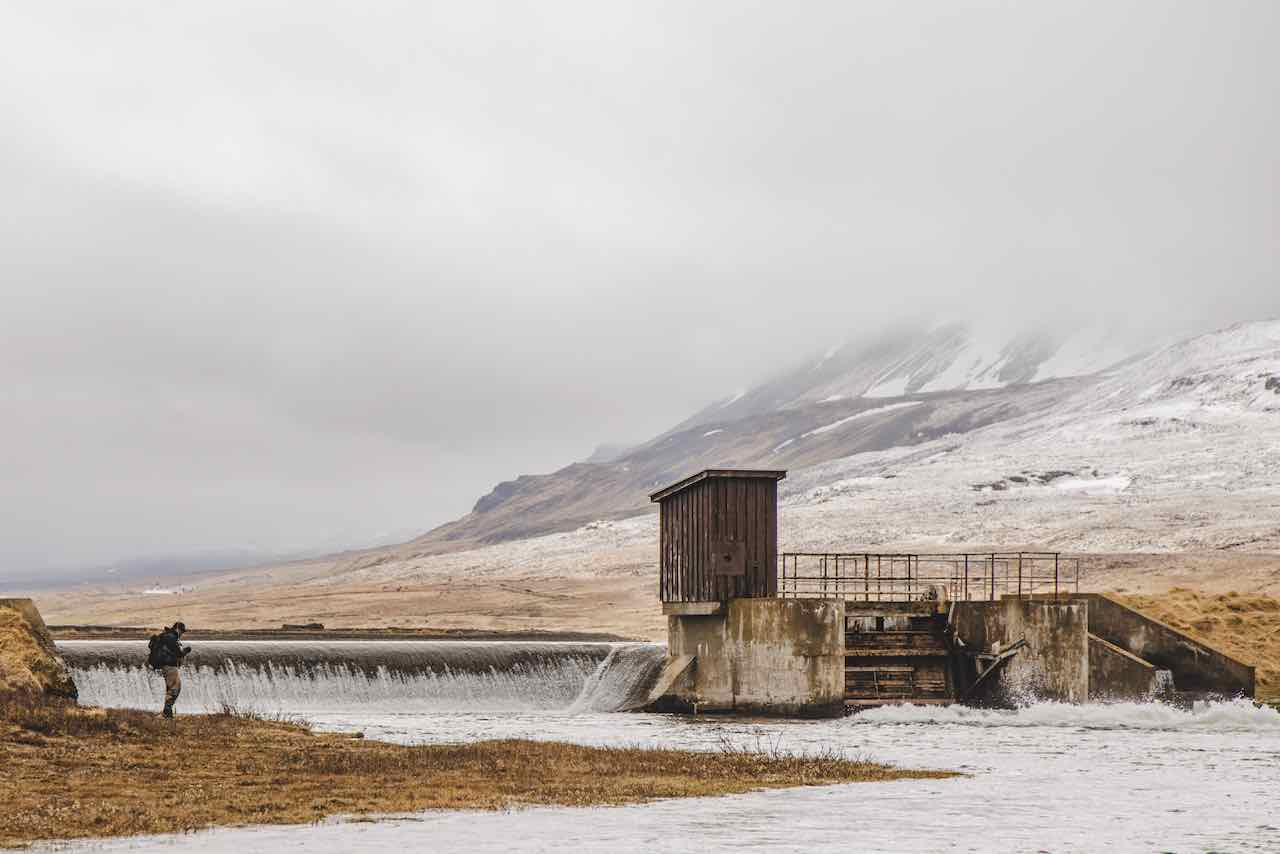 Pêcheur au bord de la rivière Laxá en Islande avec barrage et montagne enneigée en arrière plan